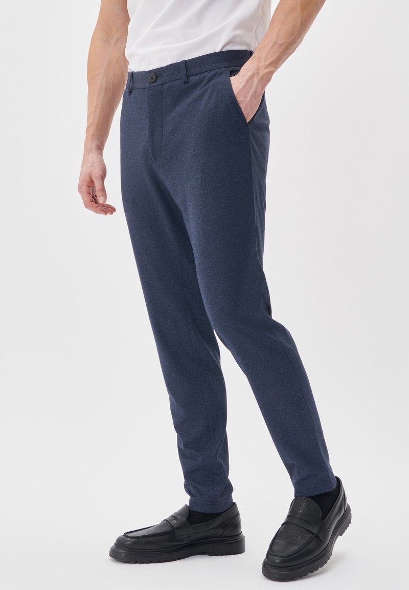 Homme portant un pantalon slim bleu marine, une chemise blanche, des chaussettes noires et des mocassins en cuir noirs, debout avec une main dans la poche.