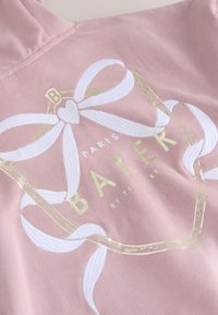 Rosa Stoff mit weißer bestickter Schleife und goldenem geometrischem Rahmen um den Text „Paris Baker by Ted Baker.“