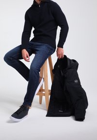 Veste noire isolante avec un col en fausse fourrure, pull côtelé bleu marine, jean bleu et baskets grises avec des accents marron, posés sur un tabouret en bois.