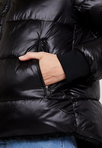 Veste matelassée noire avec une surface extérieure brillante et capitonnée. Comprend une poche zippée et des poignets en tricot côtelé, soulignant la chaleur et la fonctionnalité.