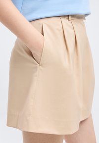 Κοντινό πλάνο ενός ατόμου που φοράει μπεζ κοντά παντελόνια με πιέτες και το χέρι του τοποθετημένο μέσα στην μπροστινή τσέπη.