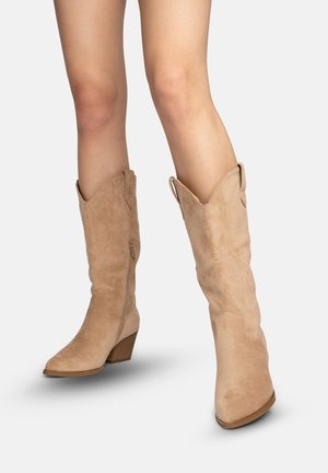 Mujer con botas vaqueras de ante color marrón claro hasta la mitad de la pantorrilla, con punta afilada y tacones bajos apilados, mostrada sobre un fondo claro y liso.