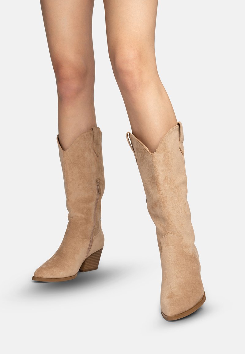 Mujer con botas vaqueras de ante color marrón claro hasta la mitad de la pantorrilla, con punta afilada y tacones bajos apilados, mostrada sobre un fondo claro y liso.