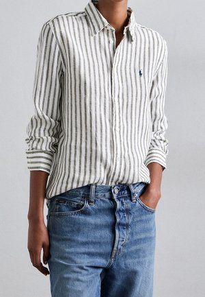 Personne portant une chemise blanche et verte à rayures verticales à manches longues rentrée dans un jean en denim bleu avec une main dans la poche.