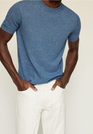 Homme portant une chemise bleue à manches courtes texturée avec les mains dans les poches d'un pantalon blanc, sur un fond uni clair.