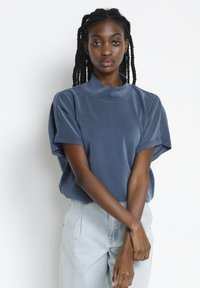 Top cu mânecă scurtă, cu guler înalt, dintr-un material moale, de culoare albastră. Prezintă o croială lejeră și o textură netedă, cu detalii subtile ale cusăturilor. Este asortat cu o pereche de pantaloni din denim deschis.