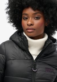 Sort jakke i puffer-stil med hætte, med en skinnende overflade, kviltet tekstur og et synligt logo på brystet. Laget over en hvid turtleneck.