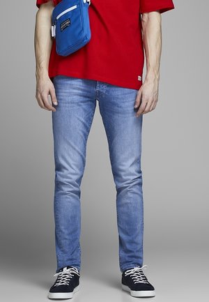 Man draagt lichtblauwe strakke jeans, zwarte sneakers met witte veters, rode shirt en een blauwe crossbody tas tegen een grijze achtergrond.