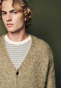 Tan, fuzzy cardigan med V-halsdesign og enkelt knaplukning, båret over en hvid og marineblå stribet skjorte. Tekstureret stof er synligt.