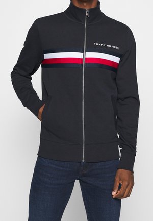 Schwarze Trainingsjacke mit Reißverschluss, weißen, roten und marineblauen horizontalen Streifen und "Tommy Hilfiger"-Logo auf der Brust, getragen mit dunkelblauer Jeans.