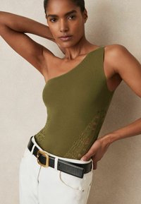 Body in maglina verde oliva con un solo spallino e inserti in pizzo sul lato. Abbinato a pantaloni bianchi e una cintura nera con accessori dorati.
