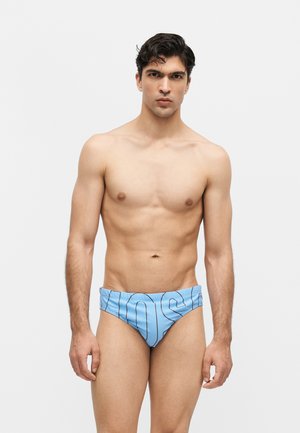 Fiatal férfi rövid fekete hajjal, világoskék fürdőnadrágot visel fekete, függőleges csíkokkal és nagy betűkkel, fehér háttér előtt áll.