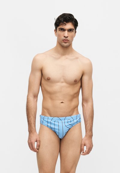 Jeune homme aux cheveux courts et noirs portant un slip de bain bleu clair avec des rayures verticales noires et de grandes lettres, debout devant un fond blanc.