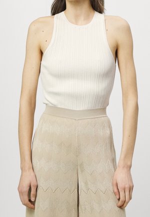Femme portant un haut en maille côtelée blanche sans manches avec un col montant et un pantalon beige texturé taille haute avec un motif en zigzag.