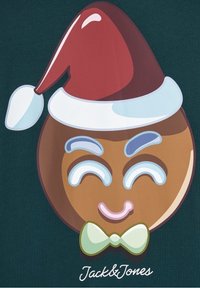 Personaje de pan de jengibre marrón con un sombrero de Santa rojo, ojos sonrientes y una pajarita verde, situado sobre un fondo verde oscuro.