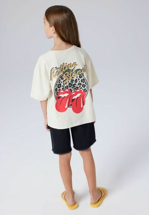Dziecko z długimi włosami, ubrane w kremowy t-shirt Rolling Stones z czerwonymi ustami i wzorem w panterkę, czarne spodenki i żółte sandały, stojące tyłem.