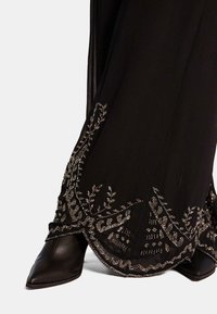 Pantaloni di tessuto neri con ricami intricati in perline argentate lungo i bordi, con scarpe con tacco nere appuntite parzialmente visibili.