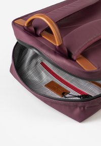 Zaino in tessuto marrone con un accento in pelle, dotato di una tasca con cerniera rossa all'interno e un rivestimento a griglia grigio.