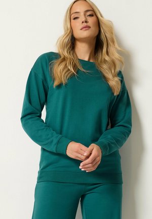 Blonde vrouw staat tegen een muur, draagt een teal kleurige trui met lange mouwen en bijpassende broek, handen losjes voor zich gevouwen.