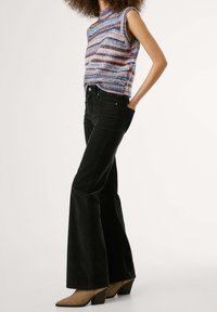 Femme portant un top en tricot rayé sans manches, un pantalon noir évasé taille haute et des bottines beige, debout avec une main dans la poche.