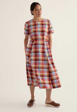 Sieviete tērpusies daudzkrāsainā pleda midi kleitā ar īsām piedurknēm un brūnās, viegli uzvelkamās koka kurpēs, stāv ar rokām kabatās.