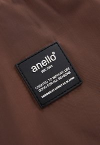 Brunt nylon tyg med en svart rektangulär etikett. Etiketten har "anello," etableringsår och en slogan i vit text.