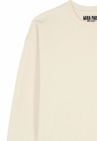 Le sweat-shirt couleur crème présente un col rond, des manches longues et une coupe décontractée. Le tissu a une texture lisse avec de subtiles taches.