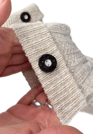 Beige gestrickter Stoff mit vertikalen Rippen, ausgestattet mit einem schwarzen Knopf mit einem weißen Loch. Eine Hand ist sichtbar, die das Kleidungsstück hält.