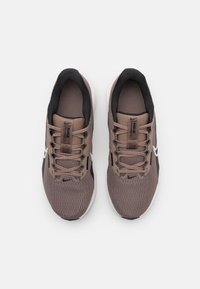 Brązowe sportowe buty z siateczki z czarnymi akcentami i białym logo w kształcie haczyka. Posiadają sznurowadła, wyściełany kołnierz oraz teksturowaną powierzchnię zapewniającą przyczepność.