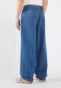 Wide-leg denim jeans in mid-blue, featuring a high waist, two back pockets, and a faded texture. Paired with gray and white sneakers.