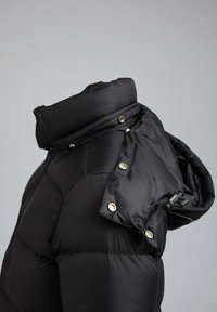 Veste matelassée noire avec un col montant, des fermetures à pression et une capuche avec cordon de serrage. Fabriquée en matière lisse et brillante avec des sections matelassées.