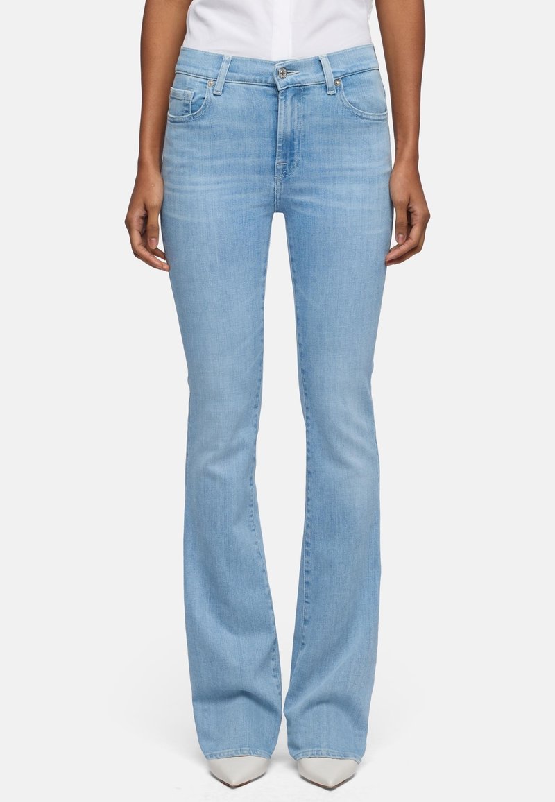 Lyseblå højtaljede bootcut-jeans båret med hvide spidse sko og en hvid top, vist fra taljen til fødderne på en neutral baggrund.