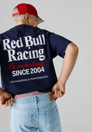 Person trägt ein navyfarbenes Red Bull Racing Shirt mit weißem und rotem Text, eine rote Kappe und hellblaue Jeans vor einem einfarbigen grauen Hintergrund.