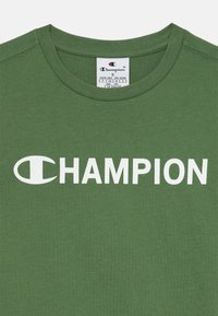 Žalias medvilninis marškinėliai su apvalia apykakle ir baltu "CHAMPION" logotipu ant krūtinės. Lygi tekstūra be matomų raštų.