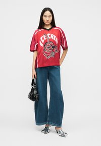 Γυναίκα που φοράει κόκκινο μακρύ πουκάμισο με σχέδιο, φαρδιά μπλε τζιν παντελόνια, ασημένια μυτερά παπούτσια και κρατάει μία μικρή μαύρη τσάντα, στέκεται σε απλό φόντο.