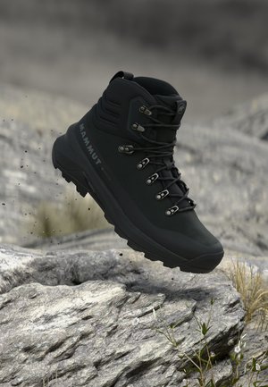 Bota de senderismo negra Mammut con ojales y cordones de metal flotando sobre un terreno rocoso con pequeñas plantas.