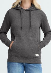 Femme portant un sweat à capuche gris foncé avec poche avant et jean bleu, mains dans les poches, étiquette de la marque visible sur l'ourlet inférieur gauche.