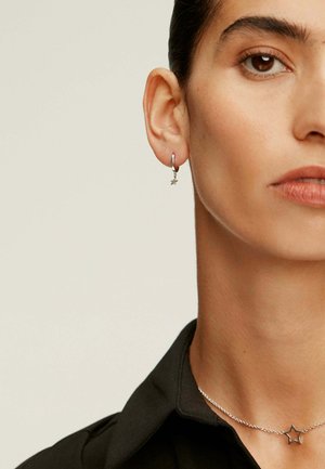 Aro de plata con un pequeño colgante de estrella y un collar de estrellas de plata en capas. El modelo lleva una camiseta negra, mostrando los detalles de la joyería.
