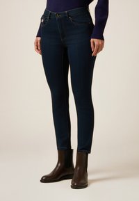 Vaqueros skinny azul oscuro con una cintura de altura media, que cuentan con bolsillos frontales curvados y detalles dorados, combinados con botas de tobillo marrones.