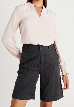 Femme portant un chemisier rose clair à manches longues avec poignets smockés et un short tailleur gris foncé à fines rayures longueur genou.