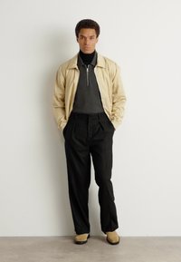 Bežs jaku ar rāvējslēdzēju virs tumša turtlenecka, apvienojot ar augsto jostasvietu melnām biksēm un gaiši brūnām kurpēm. Minimalistisks dizains, smalkas tekstūras.