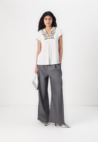 Blouse blanche brodée à manches courtes, associée à un pantalon large gris. Le mannequin tient un sac à main argenté et porte des chaussures métalliques.