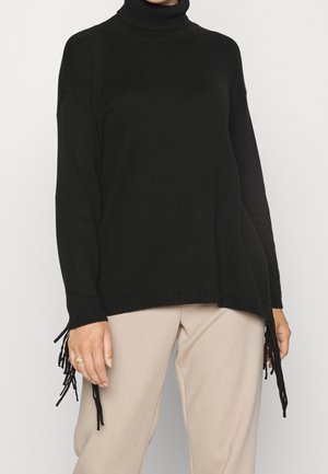 Persona che indossa un maglione nero a collo alto con dettagli di frange all'orlo e pantaloni beige su uno sfondo chiaro uniforme.