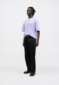 Μωβ υπερμεγεθημένη μπλούζα με ανάγλυφο λογότυπο "HUGO", συνδυασμένη με μαύρο παντελόνι cargo με τσέπες και μαύρα παπούτσια. Μινιμαλιστικό φόντο.