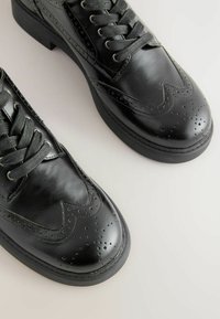 Mustat nahkakengät, joissa on brogue-muotoilu, koristeelliset rei'itykset, pyöreä kärki ja tasapohja. Sidottu yhteensopivilla mustilla kengännauhoilla.