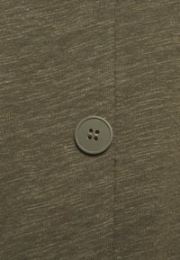 Tessuto verde scuro con finitura testurizzata, caratterizzato da un bottone rotondo grigio con due fori e una cucitura che corre verticalmente accanto a esso.