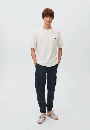 Vit t-shirt med kort ärm och en liten röd logotyp, i kombination med marinblå cargobyxor med sidofickor och dragsnöre, samt beige sneakers.