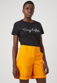 Černé bavlněné tričko s bílým logem "Tommy Hilfiger"; jasně oranžové šortky s páskem v pase a ohrnutými manžetami, vyrobené z lehkého materiálu.