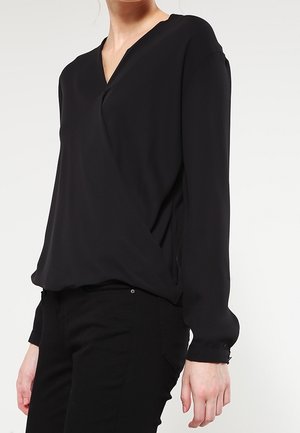 Femme portant un chemisier noir à manches longues et col en V, rentré dans un pantalon noir, debout devant un fond clair uni.