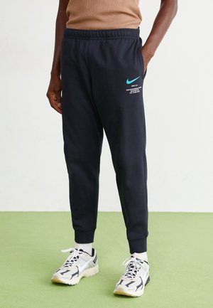 Osoba ubrana w czarne spodnie dresowe Nike z turkusowym logo w kształcie haka oraz białe buty Nike, stojąca na zielonej podłodze na tle jednolitej ściany.